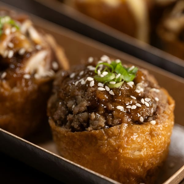 Close-up of Yum Yubu poke, a fried tofu pocket filled with ground meat, sauce, sesame seeds, and scallions.