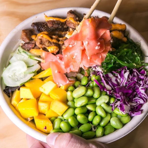 Chopsticks lifting pickled ginger from a colorful Yum Yubu poke bowl with various toppings.